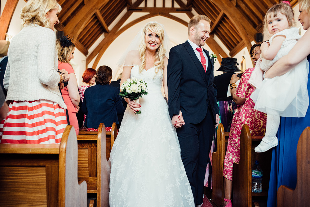Caswel House Wedding Photography Bride and groom smiling at guests in pews at church