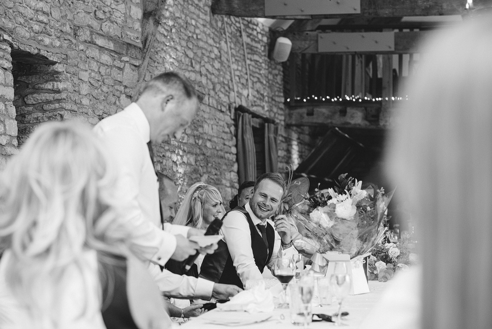 Black and white photo, groom smiling while guest gives speech