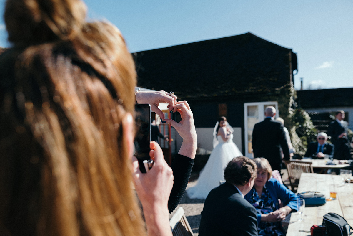 Guest taking photo of bride while second guest makes a heart shape