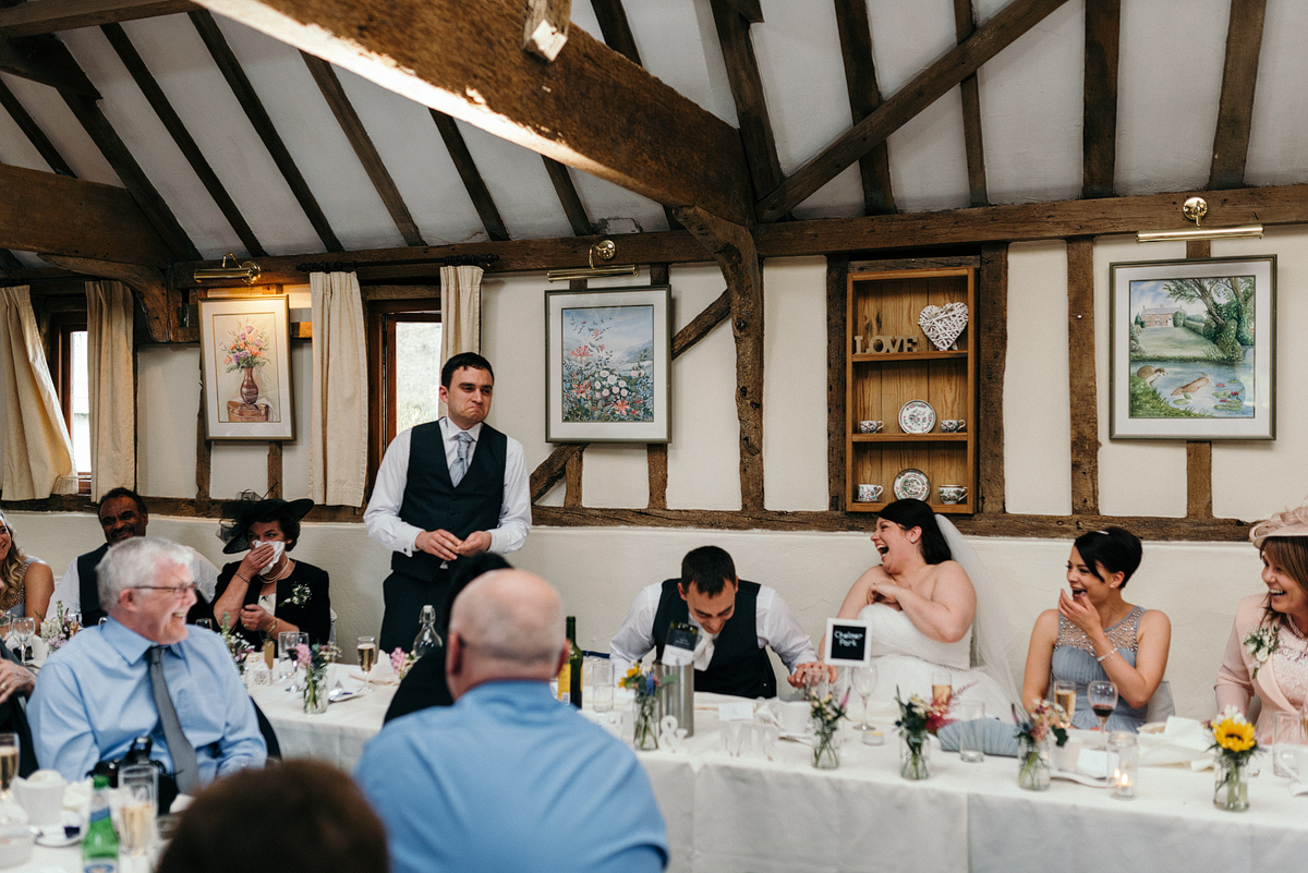 Guest makes toast as bride, groom, and guests laugh.